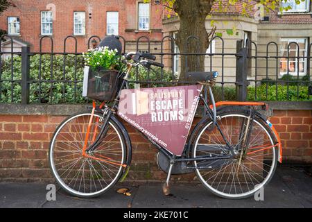 Altes Fahrrad, das an einen Metallgeländerzaun auf dem Bürgersteig gekettet wurde, der für die Werbung für ein lokales Fluchtzimmergeschäft verwendet wurde Stockfoto