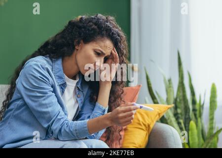 Traurige und verärgerte Frau allein zu Hause Unzufrieden mit dem Schwangerschaftstest, depressive hispanische Frau, die im Wohnzimmer auf dem Sofa sitzt. Stockfoto