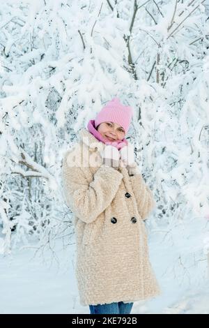 Junge Frau auf dem Hintergrund einer schönen Pflanze im Schnee. Eine junge Dame berührt bei einem Schneefall einen Ast im Schnee. Tolle Stimmung, Freude, Spaß. Stockfoto