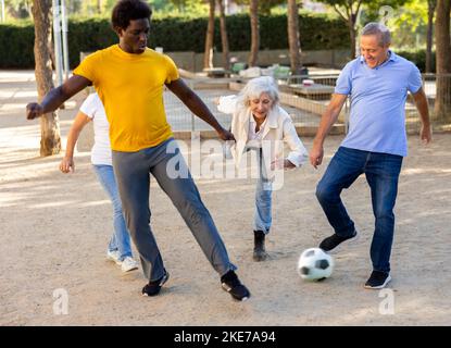 Fröhliche ältere multinationale Freunde, die an einem warmen Frühlingsabend auf einem Sandspielplatz Fußball spielen Stockfoto
