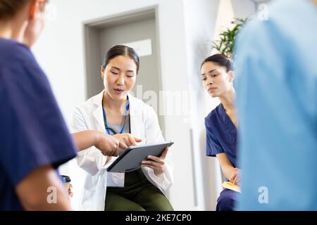 Asiatische Ärztin mit Tablette im Gespräch mit verschiedenen weiblichen Kollegen im Krankenhaus Stockfoto