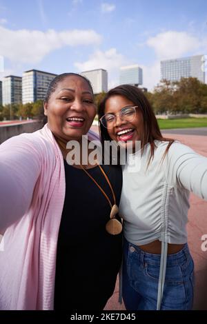 Fröhliche lateinamerikanische Mutter und Erwachsene Tochter, die fröhlich ein Selfie machen. Stadthintergrund. Stockfoto