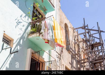 Die Kleidung trocknet im Wind auf dem Balkon eines Mehrfamilienhauses. An der Seite befindet sich ein einstürzendes Gebäude mit einem Holzgerüst. Stockfoto