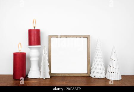 Weihnachtsmock mit Holzrahmen, weißem Baumdekor und roten Kerzen. Quadratischer Rahmen auf einem Holzregal an einer weißen Wand. Speicherplatz kopieren. Stockfoto