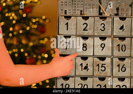 Metallischer Adventskalender im Vintage-Stil und Kinderhände in der Nähe des Weihnachtsbaums mit leuchtender Girlande auf verschwommenem Raumhintergrund. Geschenke und Überraschungen für Stockfoto