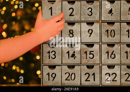 Metallischer Adventskalender im Vintage-Stil und Kinderhände in der Nähe des Weihnachtsbaums mit Spielzeugen, die Girlande auf dem Hintergrund des Zimmers leuchten. Weihnachtstraditionen und Stockfoto