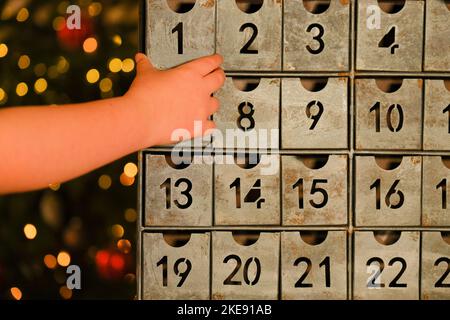 adventskalender im Vintage-Stil und Kinderhände in der Nähe des Weihnachtsbaums mit leuchtender Girlande auf verschwommenem Raumhintergrund. Weihnachtstraditionen und -Symbole. Stockfoto