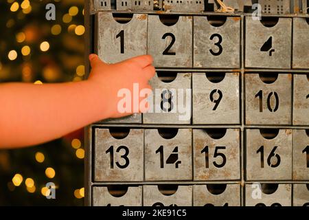 Metallischer Adventskalender im Vintage-Stil und Kinderhände in der Nähe des Weihnachtsbaums mit leuchtender Girlande auf verschwommenem Raumhintergrund. Stockfoto