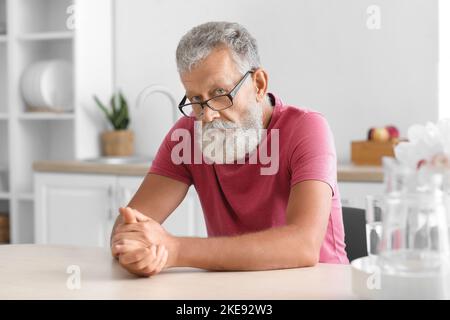 Älterer bärtiger Mann mit Brille sitzt am Tisch in der Küche Stockfoto