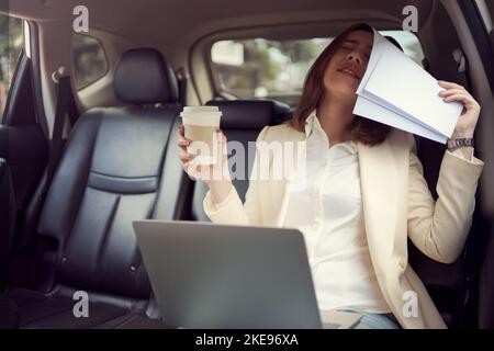 Eine junge Frau bedeckt ihr Gesicht mit einem Papierkram, während sie arbeitet und schlechte Nachrichten über ihren Job in ihrem Auto aufgibt. Stockfoto