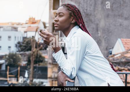 Eine Seitenansicht einer charmanten jungen afrikanischen Frau mit langen roten Zöpfen, die sich gegen den Zaun eines Café-Balkons oder ihres Hauszäunens lehnen Stockfoto