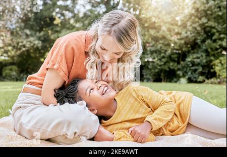 Glückliche Familie, interracial und Mädchen und Mutter entspannen sich in einem Park, glücklich und lächeln, während sie sich auf Gras ausruhen. Liebe, Familie und schwarzes Kind lachen Stockfoto