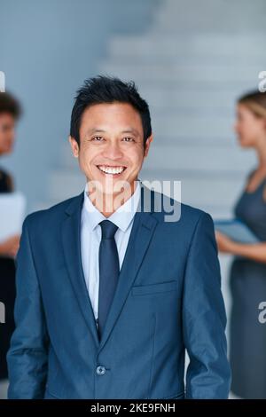 HES führt den Weg zum Erfolg. Porträt eines jungen Geschäftsmannes, der in einem Büro mit Kollegen im Hintergrund steht. Stockfoto