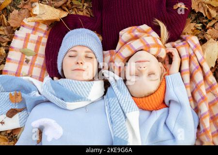 Ruhige, freundliche junge Mutter und kleine Tochter mit engen Augen in warmen blauen und orangen Outfits mit Hüten auf Laub Stockfoto