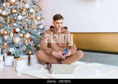 Ein Mann, der am schwarzen freitag in der Nähe des Weihnachtsbaums auf dem Boden sitzt, bestellt online Waren mit einem Laptop und einem Telefon. Glücklicher Kerl in kuscheliger Kleidung kommuniziert in sozialen Netzwerken Stockfoto