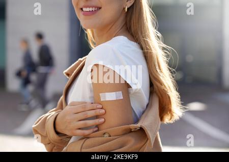 Impfkampagne von COVID-19. Junge schöne gesunde Frau, die Schulter mit Gips zeigt, Konzept der Impfung, vor dem Impfzentrum stehend Stockfoto