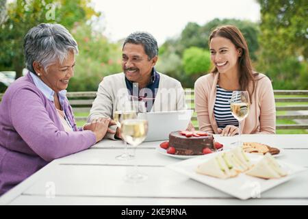 Ein inspiriertes Geburtstagsgeschenk. Ein älterer Mann zeigt seiner Familie etwas auf seinem Tablet, während sie draußen zu Mittag essen. Stockfoto