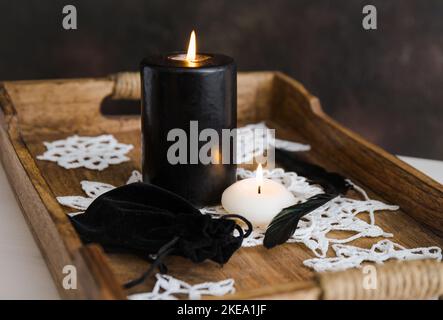 Schwarze und weiße Kerzen brennen, schwarze Samttasche mit Kordelzug, weiße handgemachte Häkelschneeflocken auf natürlichem Holztablett zu Hause, drinnen Stockfoto
