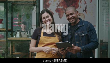 Zwei verschiedene Mitarbeiter des Coffee Shops für kleine Unternehmen. Ein schwarzer Mann hält Tablet zusammen mit einer jungen hispanischen Frau, die Schürze trägt und c anschaut Stockfoto