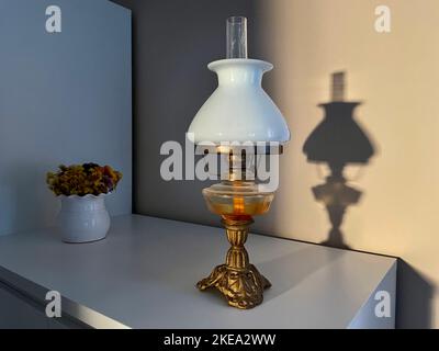 Schöne Vintage Öllampe mit Lampenschirm aus weißem Glas auf einem weißen Schrank. Schatten der Lampe an der Wand. Stockfoto