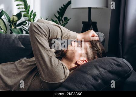 Junger gutaussehender, kranker Mann, der allein auf der Couch zu Hause liegt, an Kopfschmerzen, Krankheit, Einsamkeit, langweiligem Lebenskonzept leidet. Nahaufnahme des Reifenportraits Stockfoto