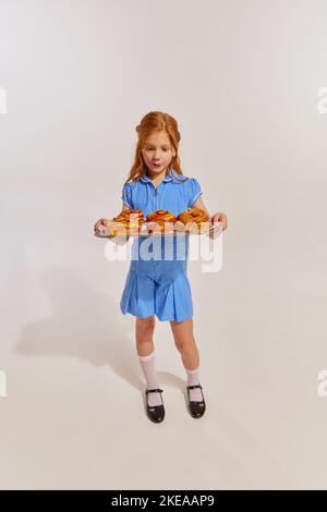 Porträt von niedlichen Rotschopf kleines Mädchen, Kind Blick auf frisch gebackene Brötchen isoliert auf grauem Hintergrund. Frühstück Stockfoto