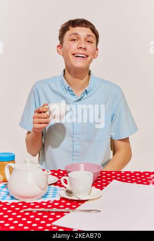 Porträt eines Jungen in blauem Hemd, der am Tisch sitzt und isoliert auf grauem Hintergrund frühstückt. Lächelnd Stockfoto