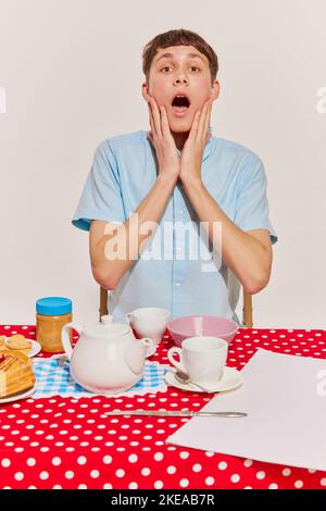 Porträt eines jungen gefühlvollen Jungen in blauem Hemd, der am Tisch sitzt und auf grauem Hintergrund isoliert frühstückt Stockfoto