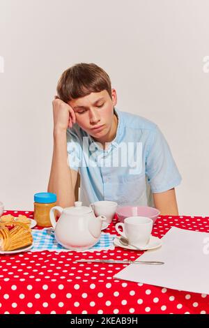 Porträt eines Jungen in blauem Hemd, der am Tisch sitzt und isoliert auf grauem Hintergrund frühstückt. Gelangweilt Stockfoto