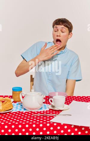 Porträt eines Jungen in blauem Hemd, der am Tisch sitzt und isoliert auf grauem Hintergrund frühstückt. Mit Tee verbrannt Stockfoto