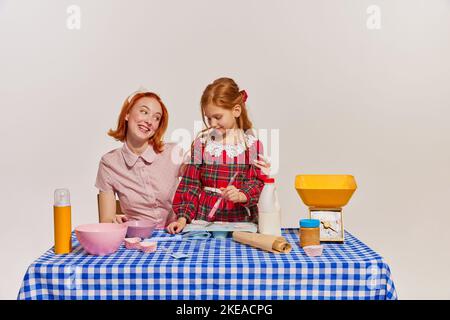 Portrait von Mutter und Tochter, die zusammen Kuchen kochen, isoliert auf grauem Hintergrund. Hobby für Mädchen Stockfoto