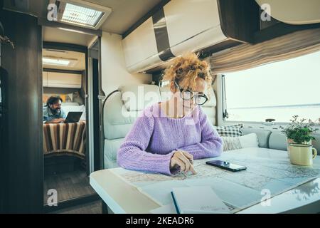 Menschen Mann und Frau im Transporter Leben Tag im Wohnmobil. Frau, die GPS-Karte des Telefons verwendet, um das nächste Ziel zu planen, und Mann, der im Hintergrund am Computer arbeitet. Stockfoto