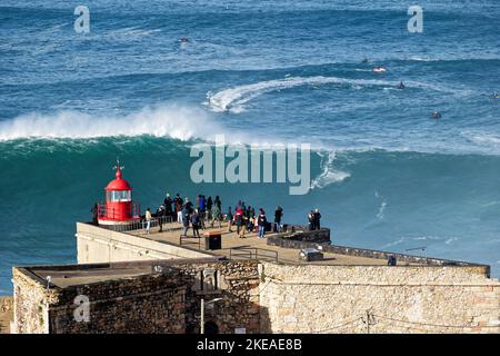 Menschen beobachten einen Surfer, der auf einer riesigen großen Welle in der Nähe des Leuchtturms Fort von Sao Miguel Arcanjo in Nazare, Portugal, reitet. Die größten Wellen der Welt. Stockfoto