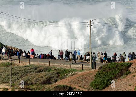 Menschen beobachten die großen riesigen Wellen, die in Nazare, Portugal, zusammenbrechen. Die größten Wellen der Welt. Touristisches Ziel zum Surfen. Stockfoto