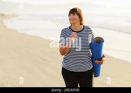 Frau im mittleren Alter, die eine Sportmatte hält und sich auf Yoga am Strand vorbereitet. Glückliche reife übergewichtige Frau, die am Meer trainiert Stockfoto