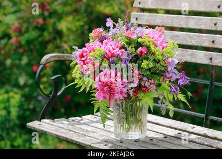 Farbenprächtiges Bouquet, rot, rosa und lila mit Pfingstrosen und Säulen in einer Glasvase auf einer hölzernen Gartenbank Stockfoto