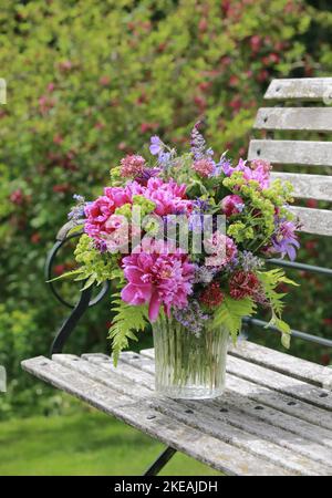 Farbenprächtiges Bouquet, rot, rosa und lila mit Pfingstrosen und Säulen in einer Glasvase auf einer hölzernen Gartenbank Stockfoto