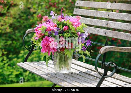 Farbenprächtiges Bouquet, rot, rosa und lila mit Pfingstrosen und Säulen in einer Glasvase auf einer hölzernen Gartenbank Stockfoto