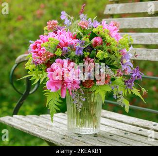 Farbenprächtiges Bouquet, rot, rosa und lila mit Pfingstrosen und Säulen in einer Glasvase auf einer hölzernen Gartenbank Stockfoto