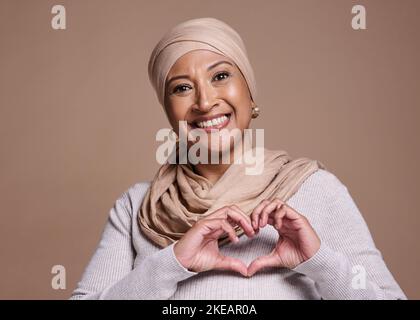 Hände, Herz und muslim mit einer reifen Frau im Studio auf braunem Hintergrund, um Liebe oder Gesundheit zu fördern. Porträt, Handzeichen und Islam mit einem Senior Stockfoto