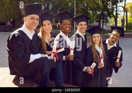 Junge fröhliche Menschen graduieren an der Universität in schwarzem Gewand und stehen auf der Straße Stockfoto