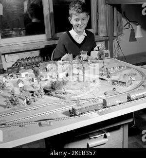 Junge in den 1950s. Ein Junge an seiner Modelleisenbahn, montiert auf einem Tisch in seinem Zimmer. Er leitet die Züge, wie schnell es geht und ob er will, dass der Zug vorwärts oder rückwärts fährt. Die Modelleisenbahn hat Häuser, Bäume, Tiere und Menschen in ihr als maßstabsgetrete Modelle. Höchstwahrscheinlich ein Zugsatz des deutschen Herstellers Fleischmann, da die Schienen das typische zweigleisige Gleichstrom-System sind. Schweden 1958. Conard Ref. 3867 Stockfoto