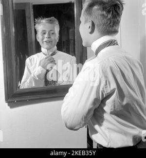 In der 1950s anziehen. Ein junger Mann vor dem Spiegel knotet seine Krawatte. Es gibt Varianten der Bindung der Krawatte einschließlich der Ascot, Schleife, Bolo, Reißverschluss Krawatte, Krawatte und Strick. Zu der Zeit, als das Bild aufgenommen wurde, waren Mode und Aussehen der Krawatten schlanker und dünner als frühere Jahrzehnte. Es gibt vier Hauptknoten, die verwendet werden, um Krawatten zu Knoten; vier-in-Hand-Knoten, Pratt-Knoten, Half-Windsor-Knoten und der Windsor-Knoten. Schweden 1959. Conard Ref. 4019 Stockfoto