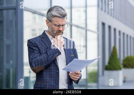 Reifer grauhaariger Geschäftsmann, der den Finanzbericht denkt und liest, Senior man im Geschäftsanzug vor dem Bürogebäude mit Rechnungen. Stockfoto