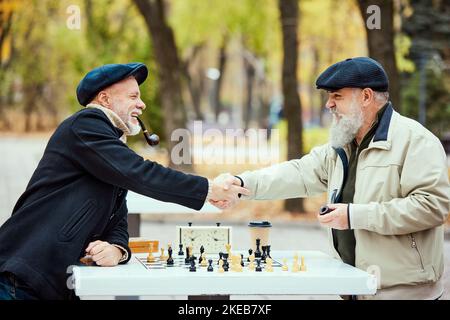 Porträt von zwei älteren Männern, die im Park an einem Herbsttagaus Schach spielen. Gutes Spiel. Händeschütteln. Konzept der Freizeitgestaltung, alte Generation Stockfoto