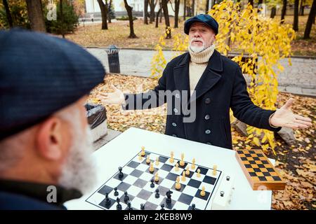 Porträt von zwei älteren Männern, die im Park an einem Herbsttagaus Schach spielen. Hände verteilen. Gewinnen. Konzept der Freizeitgestaltung, alte Generation Stockfoto