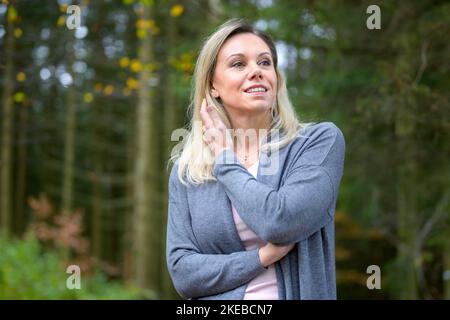 Die wunderschöne, sanft lächelnde blonde Frau mittleren Alters läuft ihre Hand durch ihre Haare im Wald Stockfoto