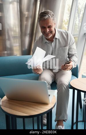 Ein fröhlicher Mann mittleren Alters sitzt auf dem Sofa in einem Erholungsgebiet Stockfoto