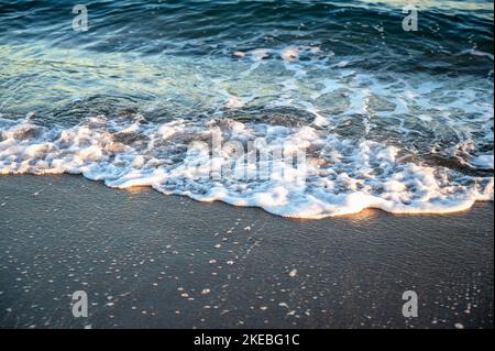 Blaues und weißes Meerwasser auf nassem Strandsand bei Sonnenaufgang. Stockfoto