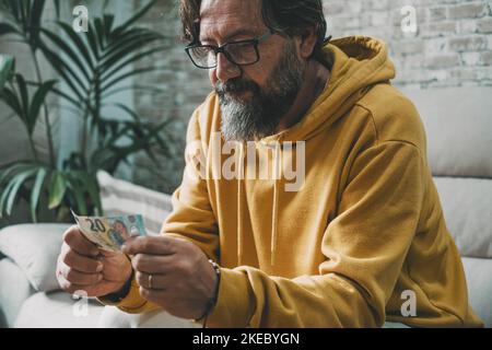 Wirtschaft und Geld Menschen Problem Konzept. Mann mit Geldschein und besorgter Gesichtsausdruck. Reifer kaukasischer Mann, der zuletzt 20 Euro auf dem Sofa sitzt. Real Life Cash und Rechnungszahlungen Stockfoto